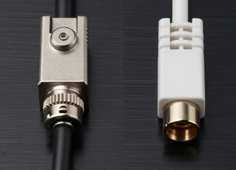 A side-by-side comparison of a heavy-duty military-grade optical connector and a standard commercial fiber optic connector on a metallic surface.